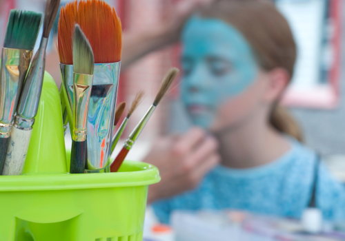 A close-up of paintbrushes in a green holder, with a child in the background getting her face painted blue.