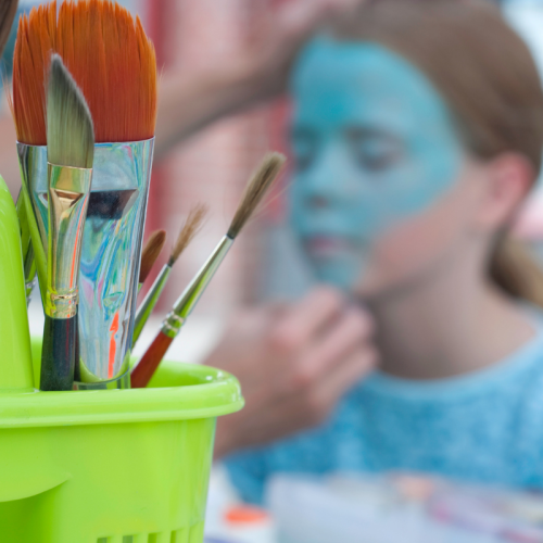 A close-up of paintbrushes in a green holder, with a child in the background getting her face painted blue.