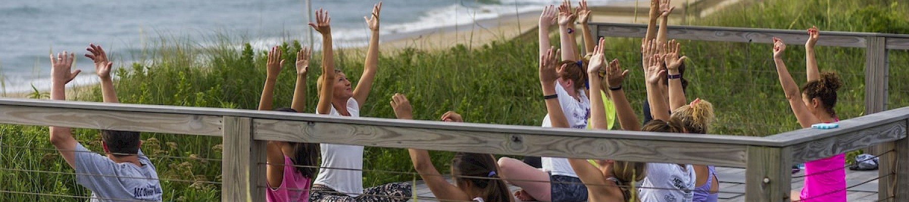 A group of people is practicing yoga on a wooden deck, stretching with raised arms, alongside an ocean shoreline, enjoying the scenic view.