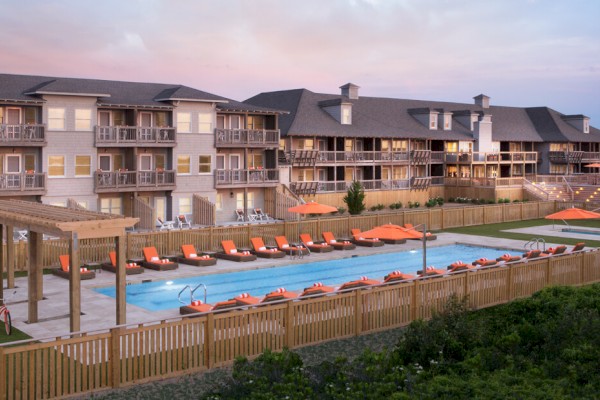 The image shows a residential complex with balconies and a large outdoor pool area. There are lounge chairs, umbrellas, and a pergola beside the pool.