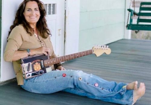A woman sitting on a porch, smiling, holding a guitar made from a cigar box, while wearing jeans with colorful buttons.