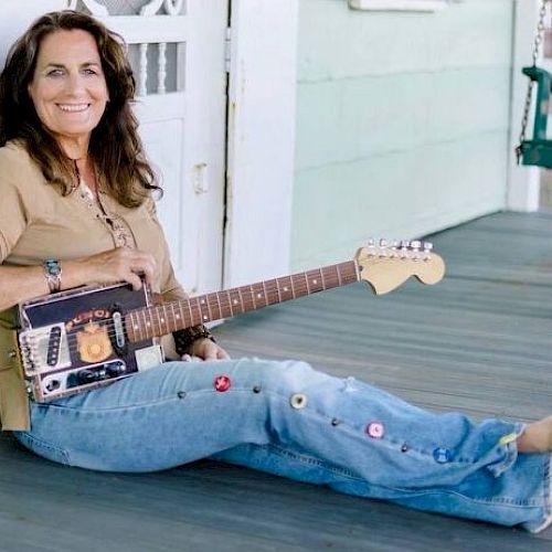 A woman sitting on a porch, smiling, holding a guitar made from a cigar box, while wearing jeans with colorful buttons.