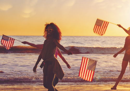 People are walking on a beach at sunset, holding American flags, with waves in the background and a sky filled with soft clouds.