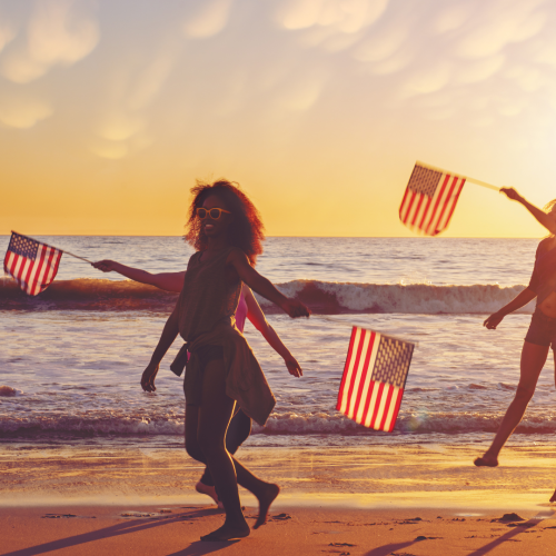 People are walking on a beach at sunset, holding American flags, with waves in the background and a sky filled with soft clouds.