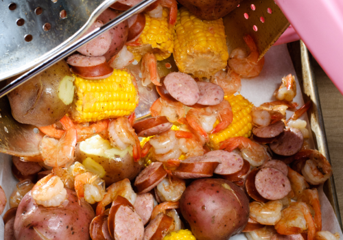 The image shows a seafood boil being poured out of a pot. Mixed in are shrimps, sausages, corn on the cob, and red potatoes on a tray.