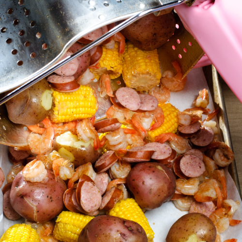 The image shows a seafood boil being poured out of a pot. Mixed in are shrimps, sausages, corn on the cob, and red potatoes on a tray.