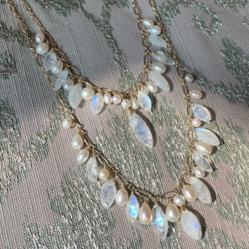 The image shows two gold chain necklaces adorned with pearls and iridescent gemstone beads, placed on a textured surface with a floral pattern.