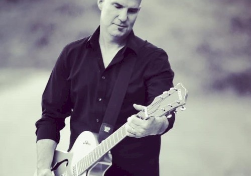A person is playing an electric guitar outdoors, wearing a dark shirt. The background is blurred, and the image is in black and white.