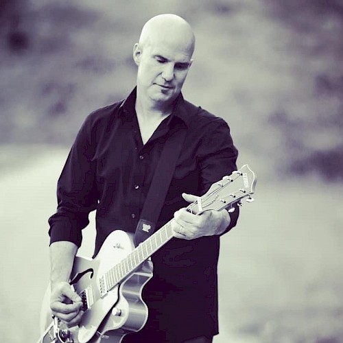 A person is playing an electric guitar outdoors, wearing a dark shirt. The background is blurred, and the image is in black and white.