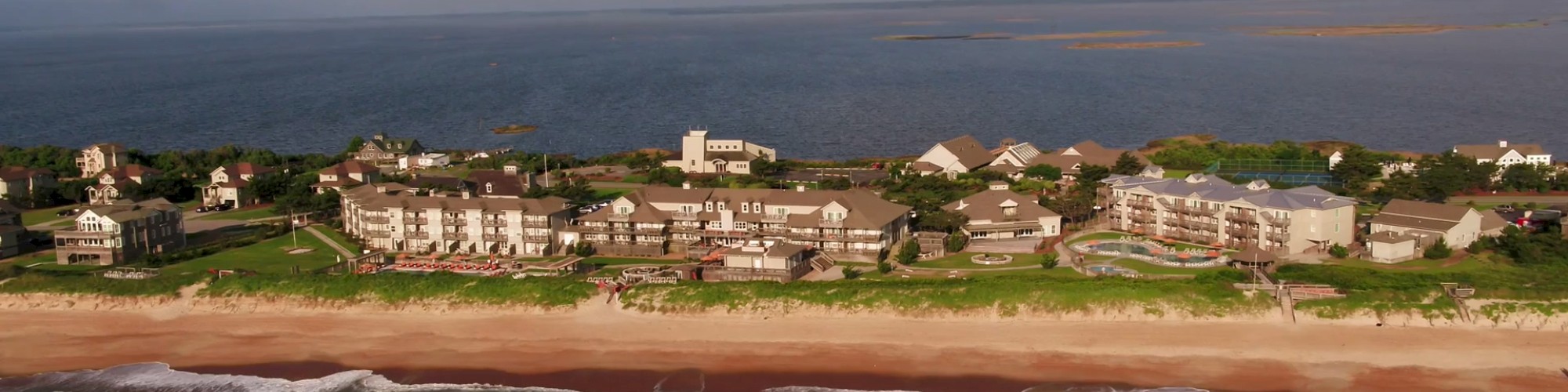 Aerial view of a beachside resort area with buildings and houses, situated along the coastline with the ocean in the background and a cloudy sky.