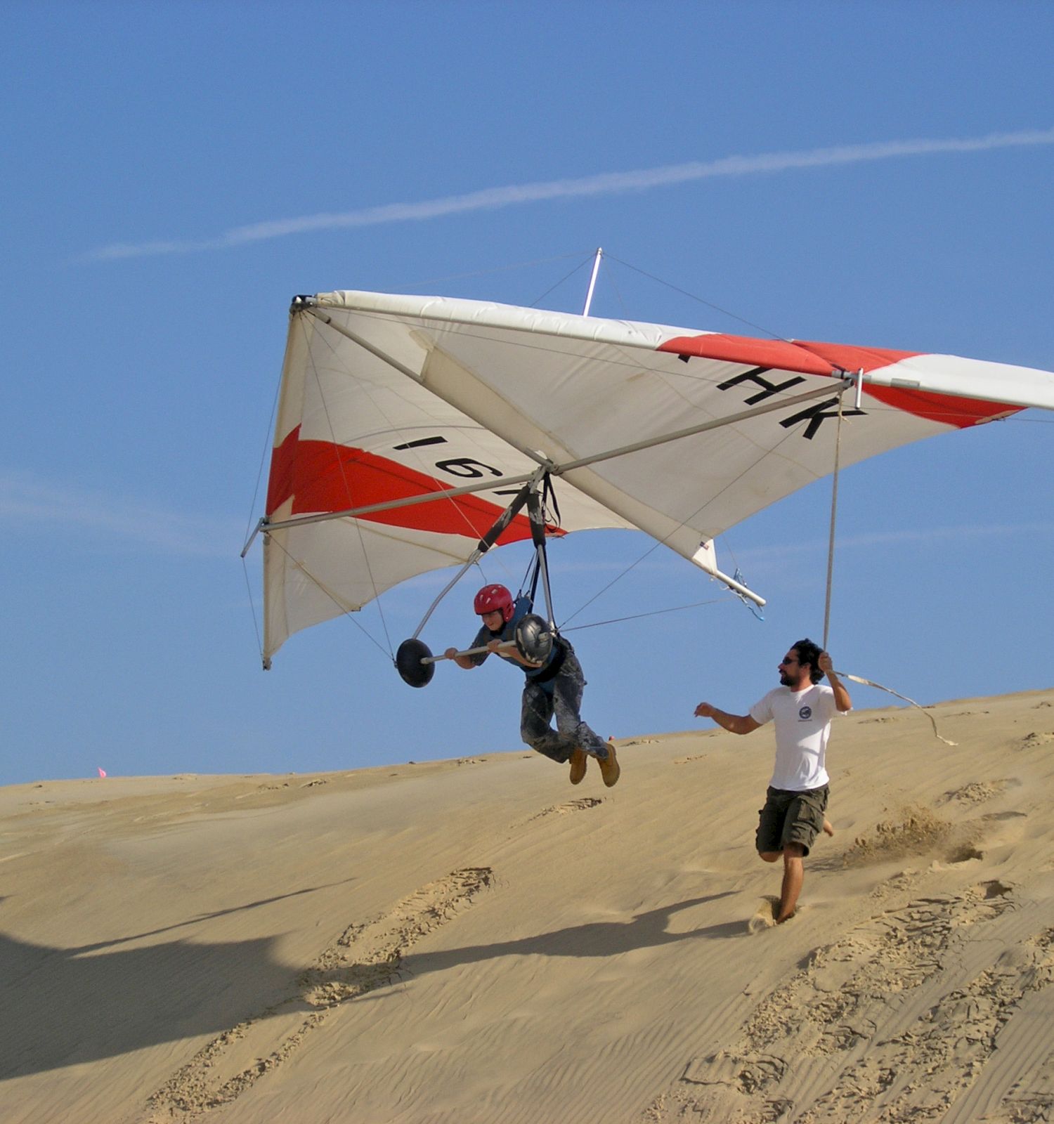 A person is hang gliding over a sandy area while another individual on the ground appears to be either assisting or watching them take off.