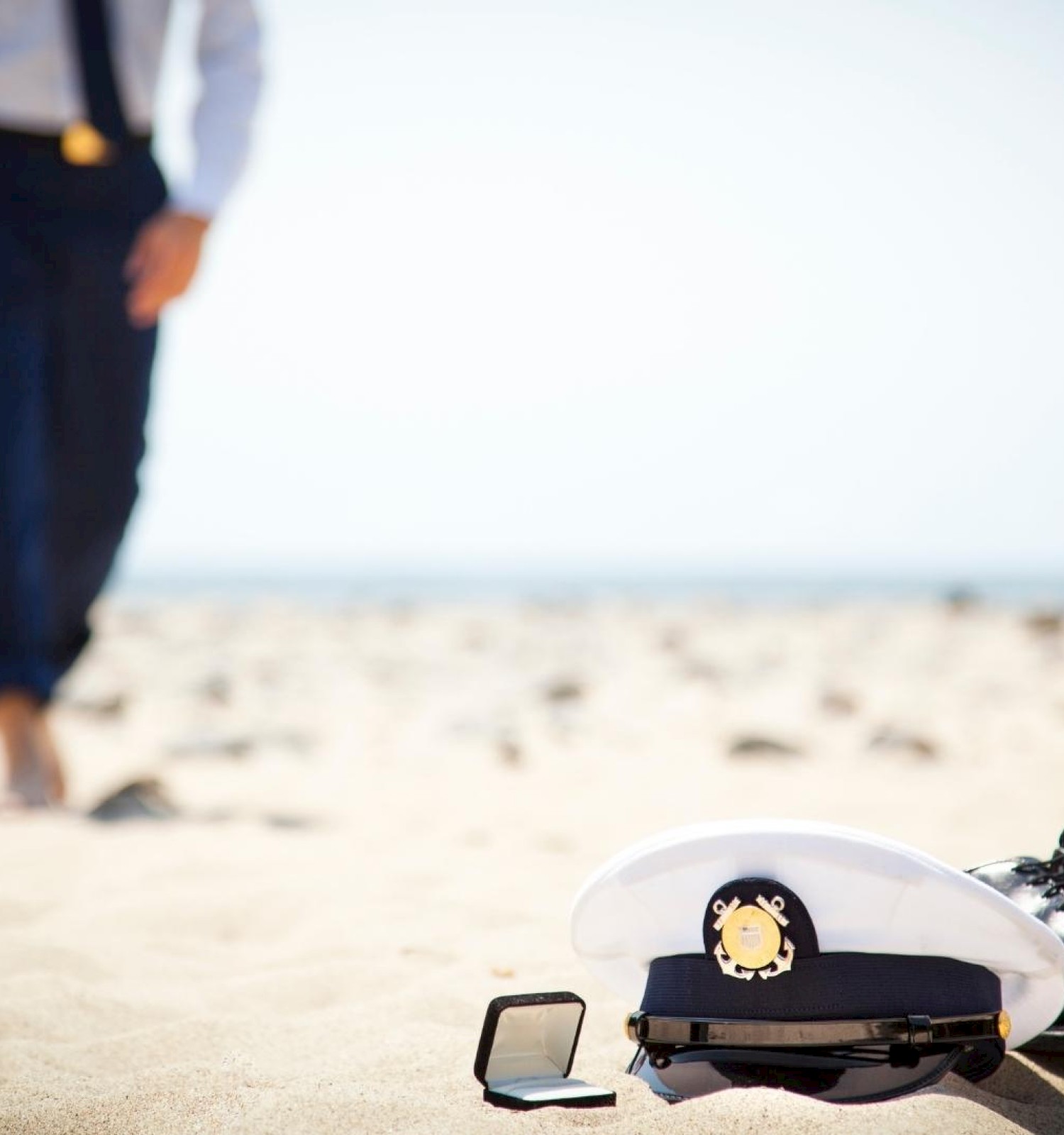 A couple holding hands walks on a sandy beach; foreground shows a marine officer's cap, belt, and shoes laid on the sand.