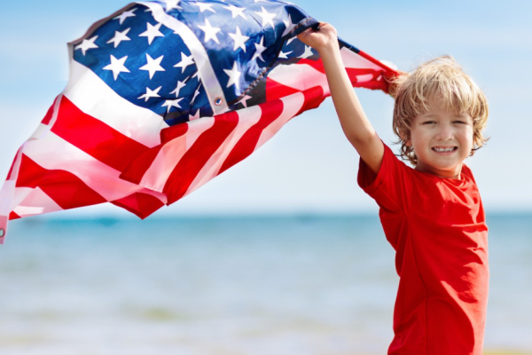 A child at the beach holds an American flag, wearing red, white, and blue clothing, smiling with the sea and sky in the background.