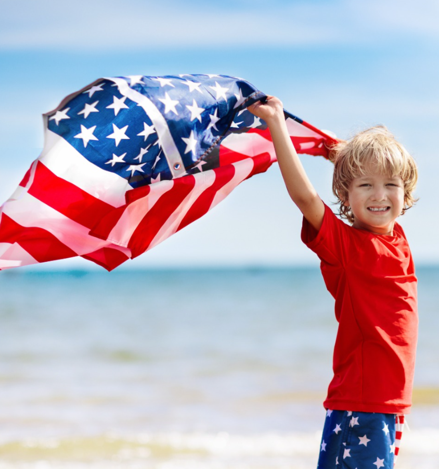 A child at the beach holds an American flag, smiling and wearing patriotic clothing, with the ocean and a clear sky in the background.