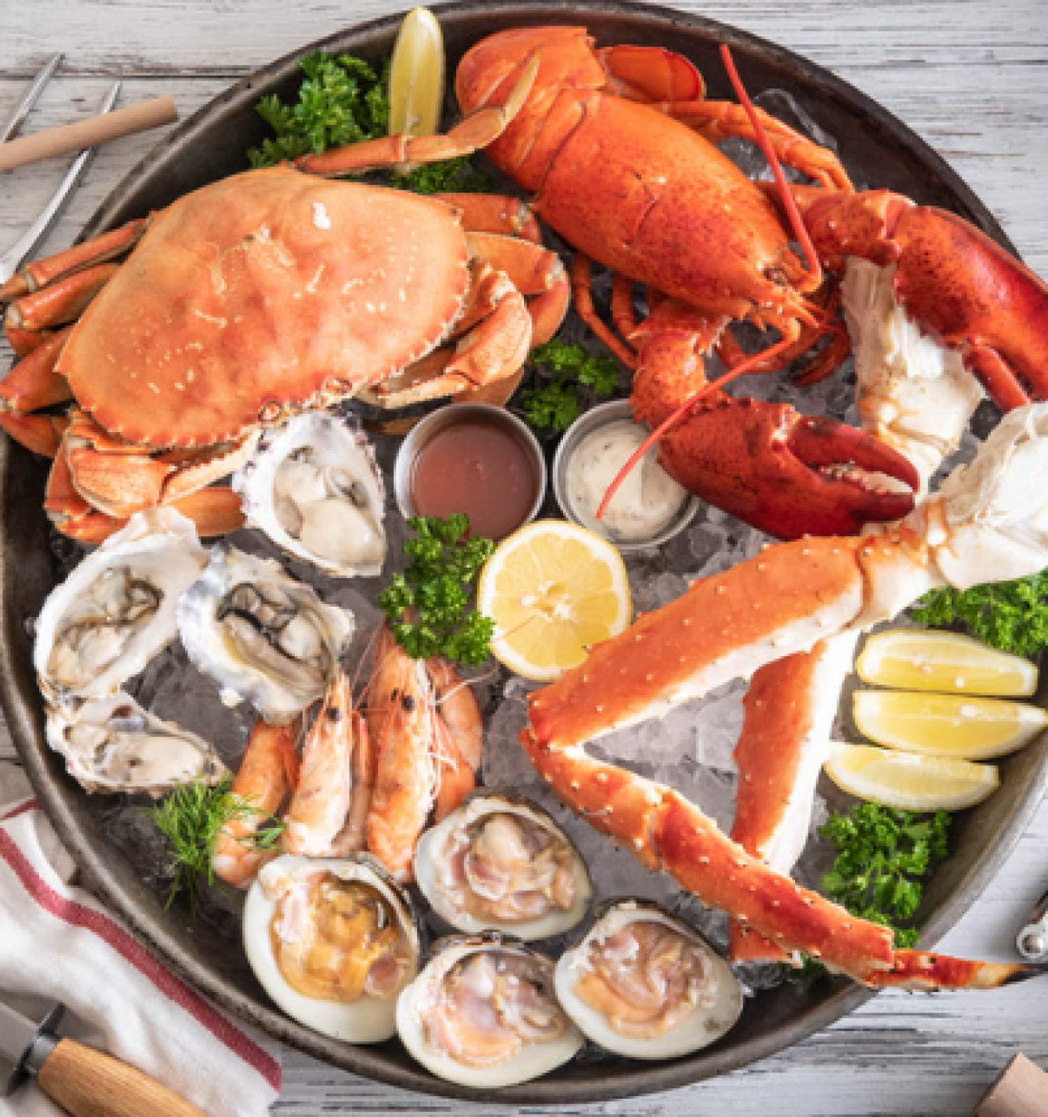 The image shows a seafood platter with crab, lobster, oysters, shrimp, lemon wedges, and sauces, presented on ice with tools for cracking shells.