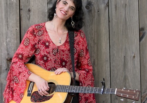 A woman in a red patterned blouse smiles while holding an acoustic guitar, standing against a wooden backdrop, wearing dangling earrings.