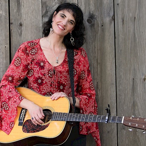 A woman in a red patterned blouse smiles while holding an acoustic guitar, standing against a wooden backdrop, wearing dangling earrings.