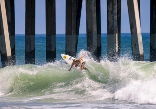 A surfer is performing a maneuver on a wave near a pier, with the ocean and pier supporting pylons in the background.