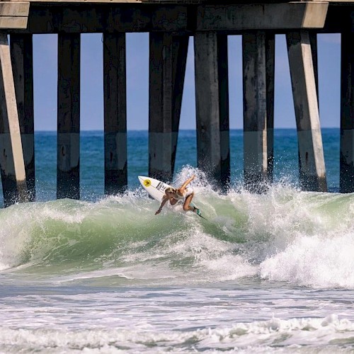 A surfer is performing a maneuver on a wave near a pier, with the ocean and pier supporting pylons in the background.