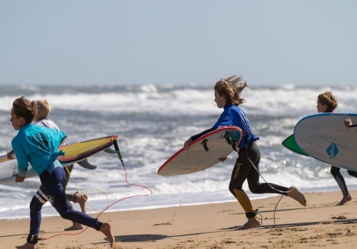 Four people are running on a sandy beach toward the ocean, each carrying a surfboard, dressed in wetsuits.