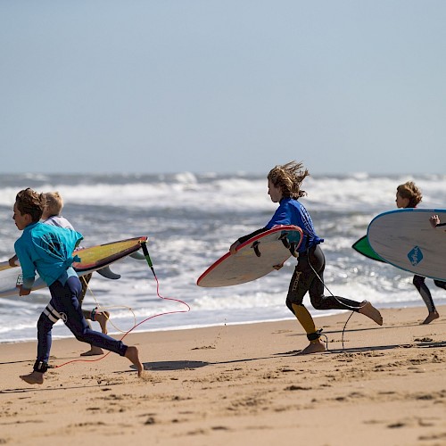 Four people are running on a sandy beach toward the ocean, each carrying a surfboard, dressed in wetsuits.