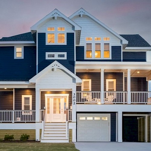 A three-story house with a white and dark blue exterior, large windows, multiple balconies, and a garage, set against a twilight sky.