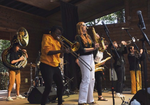 A band performs on an outdoor stage with brass and woodwind instruments, and a drummer in the background, setting an energetic atmosphere.