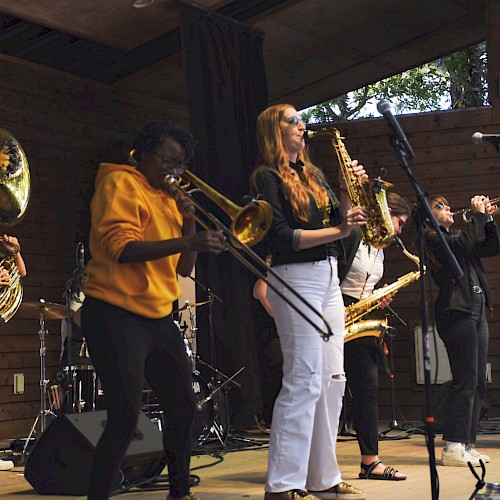 A band performs on an outdoor stage with brass and woodwind instruments, and a drummer in the background, setting an energetic atmosphere.