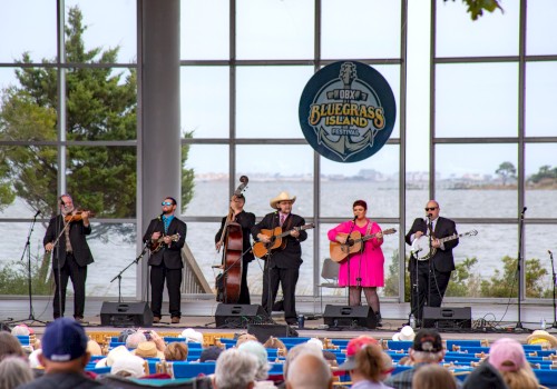 A band performs on stage with a Bluegrass Island sign in the background, as an audience watches by the water.