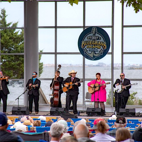 A band performs on stage with a Bluegrass Island sign in the background, as an audience watches by the water.