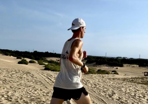 A person is running on sandy terrain wearing a white tank top, black shorts, and a white cap. The background shows a clear sky and some vegetation.