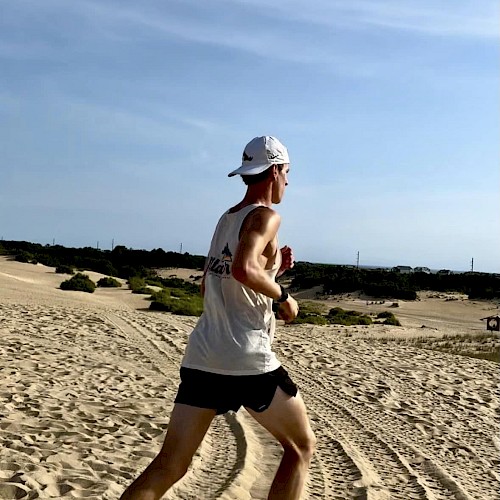 A person is running on sandy terrain wearing a white tank top, black shorts, and a white cap. The background shows a clear sky and some vegetation.