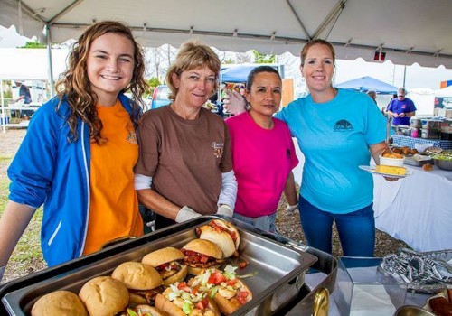 Four women are standing together and smiling under a tent. In front of them are trays filled with food, including sandwiches and other dishes.