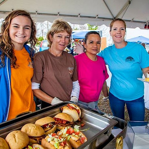 Four women are standing together and smiling under a tent. In front of them are trays filled with food, including sandwiches and other dishes.