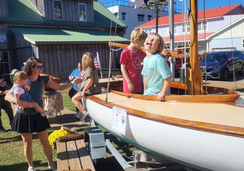 People, including children, are gathered around a wooden sailboat on display outdoors. Yellow flowers and a building are in the background.