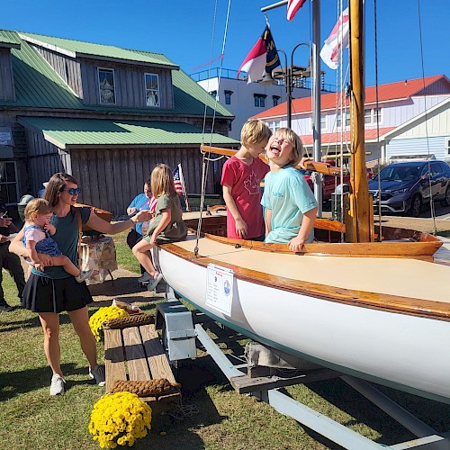 People, including children, are gathered around a wooden sailboat on display outdoors. Yellow flowers and a building are in the background.