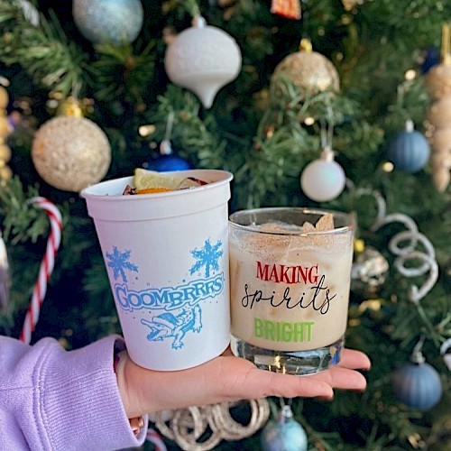 A person holding two holiday-themed drinks in front of a decorated Christmas tree with ornaments and candy canes.