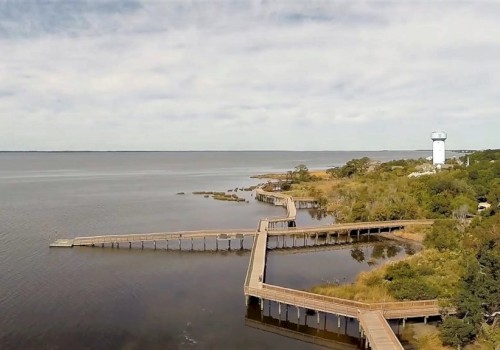 A scenic coastal view features a long, winding boardwalk over the water, leading to a lighthouse surrounded by greenery.