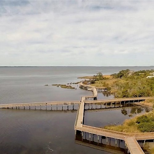 A scenic coastal view features a long, winding boardwalk over the water, leading to a lighthouse surrounded by greenery.