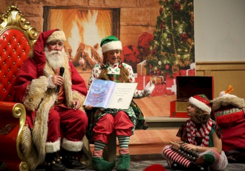 Santa Claus and two elves sit by a fireplace with a Christmas tree, as one elf reads a book.