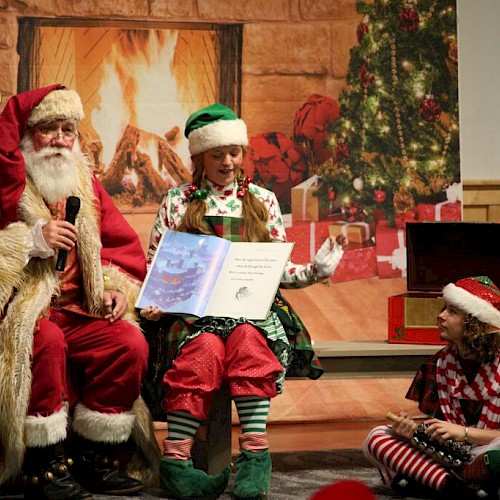 Santa Claus and two elves sit by a fireplace with a Christmas tree, as one elf reads a book.