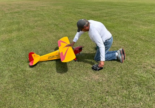 A person kneels on grass, adjusting a yellow and red model airplane in preparation for flight, while holding a remote control.