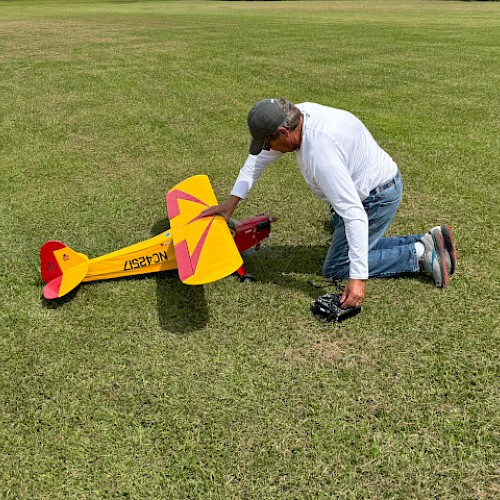 A person kneels on grass, adjusting a yellow and red model airplane in preparation for flight, while holding a remote control.