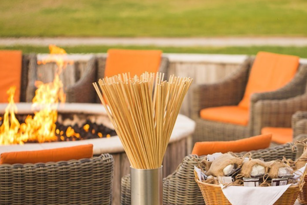 A cozy outdoor setting with orange-cushioned wicker chairs, a fire pit, and a basket of supplies for s'mores, featuring marshmallow skewers.