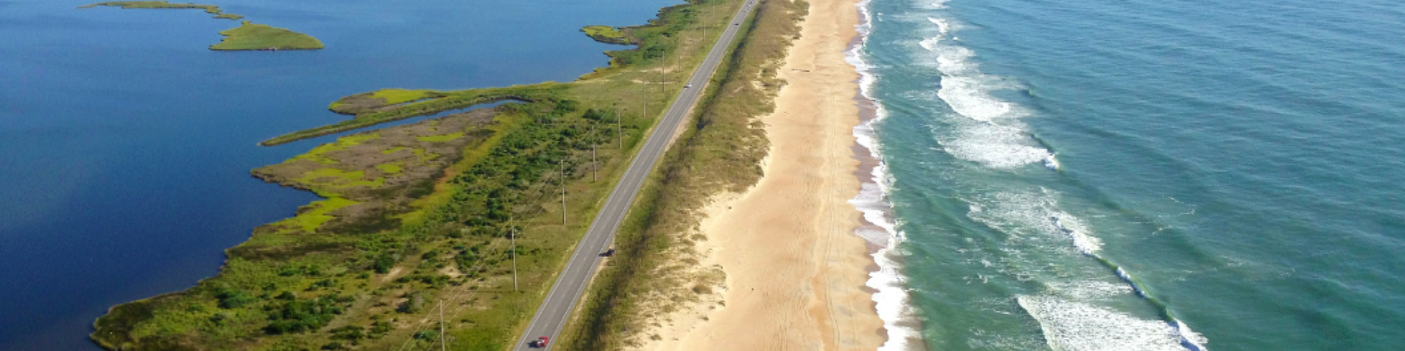 An aerial view of a coastline with a road parallel to a sandy beach on one side and a body of water on the other side, under a clear blue sky.