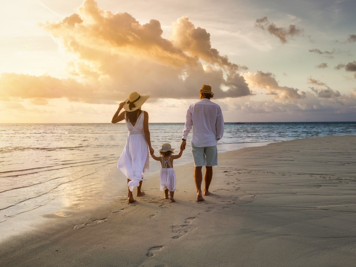 A family of three walks hand in hand on a beach at sunset, wearing light clothing and hats, with footprints in the sand behind them.