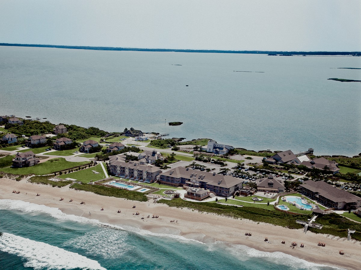 An aerial view shows a coastal resort, with buildings, a pool, and a beach along the shoreline, leading out to the ocean.