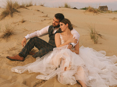 A couple in formal attire sits closely on sand dunes, surrounded by grasses, under a soft sky, creating a serene, romantic scene.