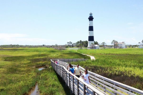 A group of people walk on a wooden path through grassy wetlands, with a black and white lighthouse in the background under a clear blue sky.