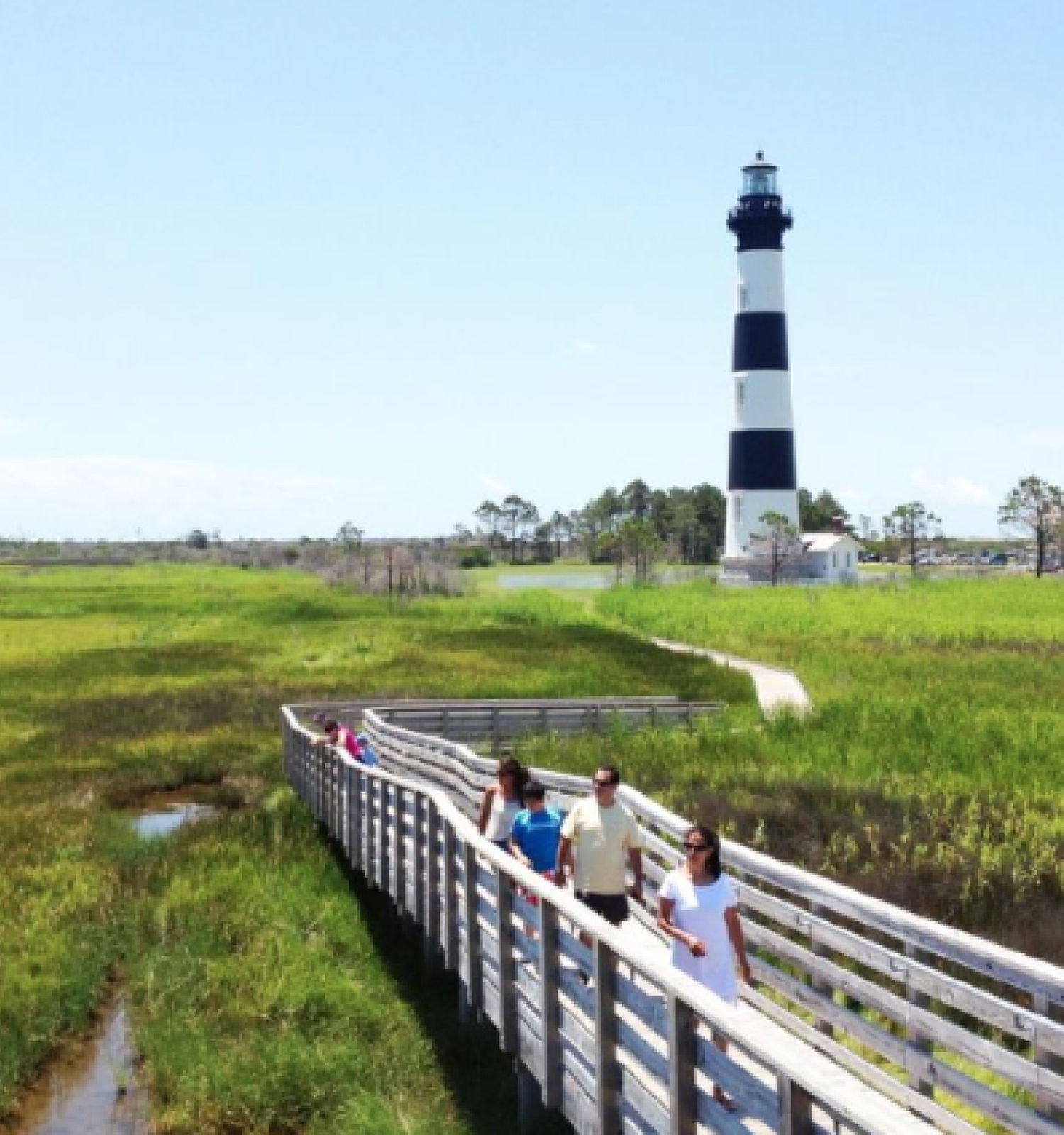 A group of people walk along a boardwalk through a grassy landscape, with a black and white striped lighthouse in the background.